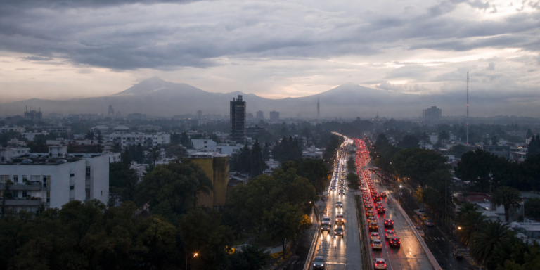 Tráfico vehicular en una avenida de la Ciudad de México al amanecer, con pavimento mojado y cielo nublado.